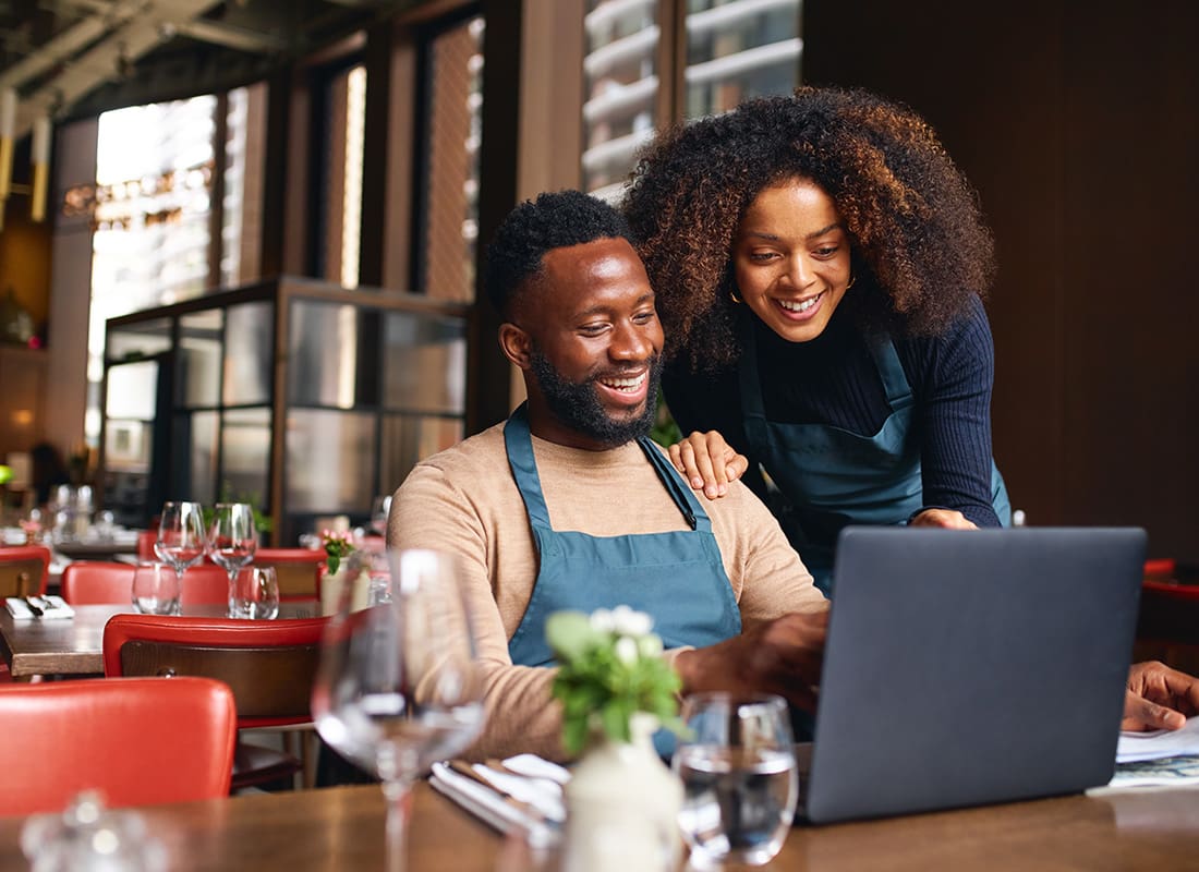 Business Insurance - Portrait of Two Cheerful Restaurant Managers Sitting at a Table While Using a Laptop
