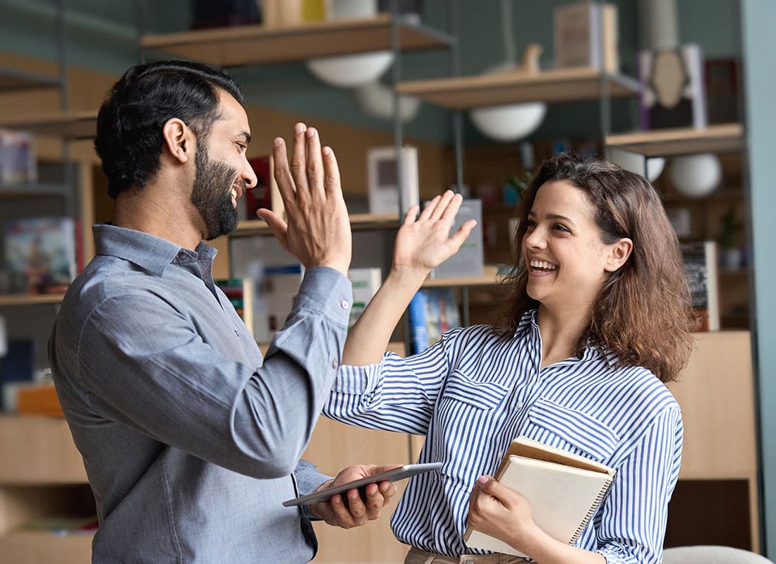 Employee Benefits - Two Cheerful Young Diverse Employees Giving Each Other High Fives While Standing in the Office